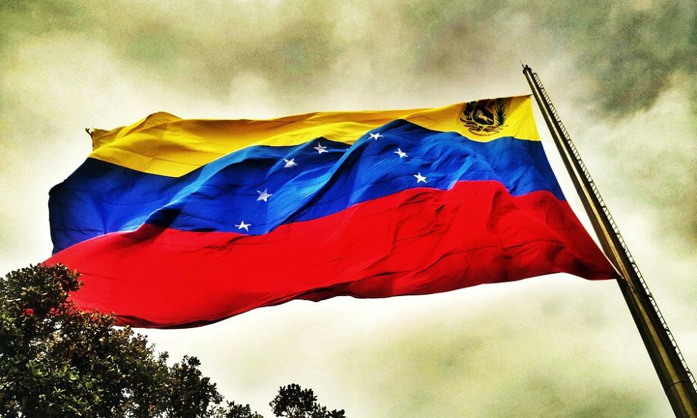 Bandera de Venezuela ondeando con fuerza sobre cielo nublado y árboles